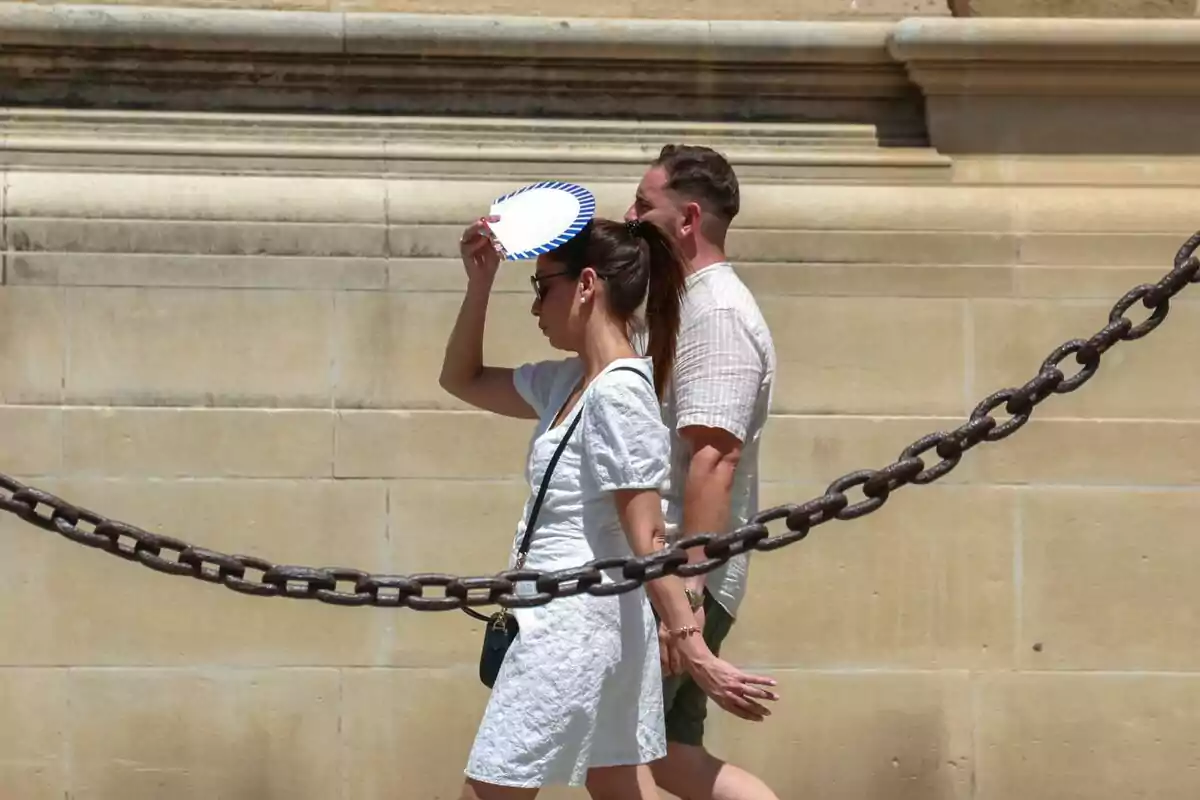 Dues persones caminen al costat d’una paret de pedra mentre una d’elles es protegeix del sol amb un ventall.