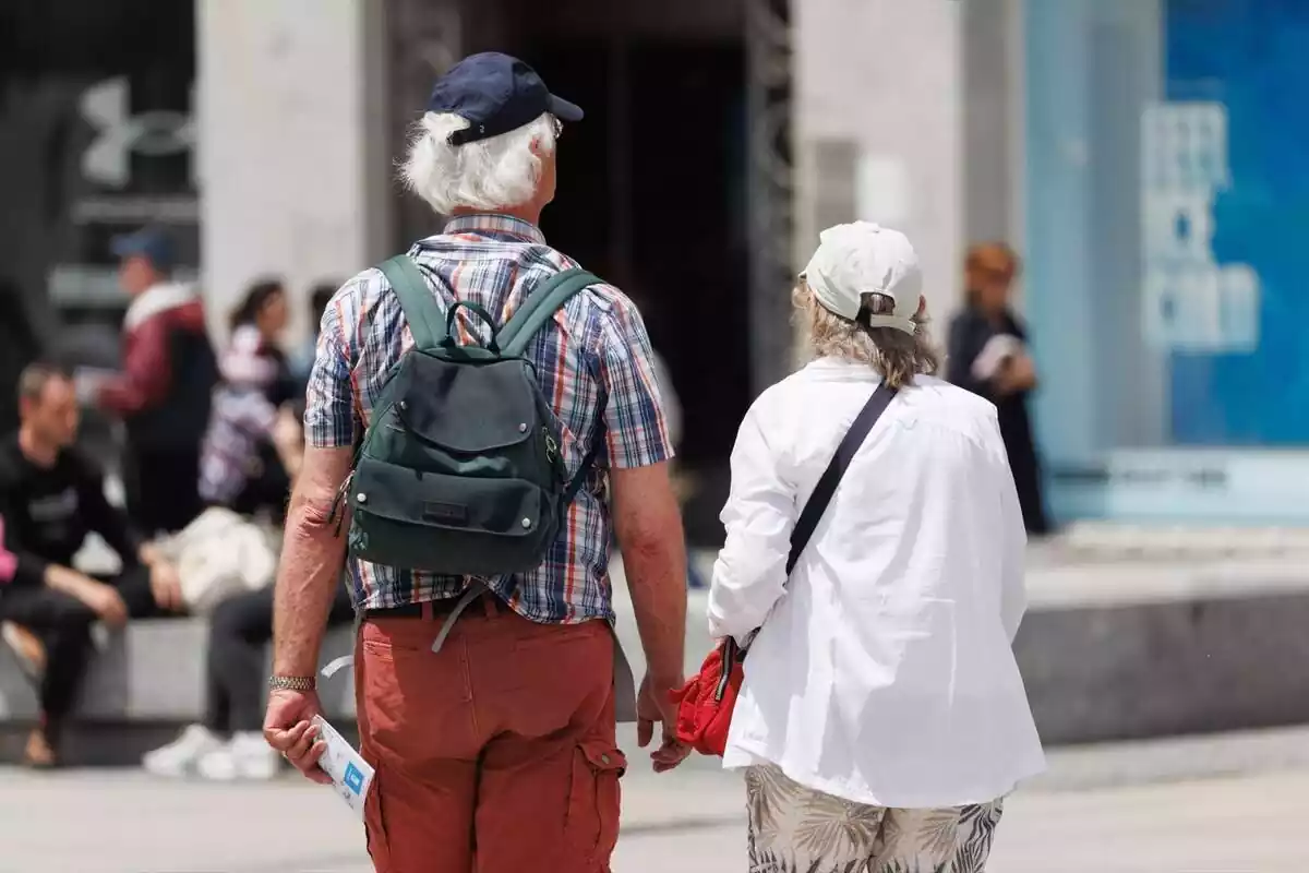 Una parella de jubilats d'esquena, amb gorra i motxilla, caminant pel carrer