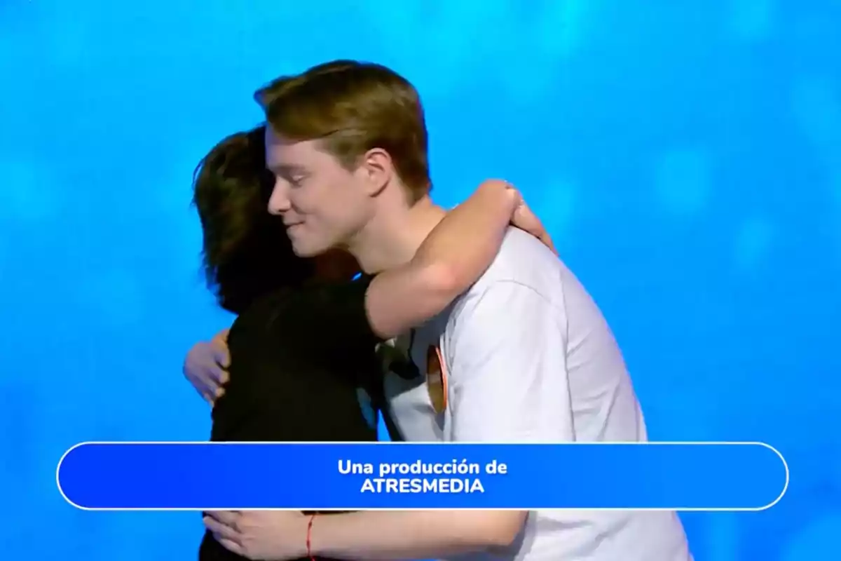Rosa and Manu hug in front of a blue background with a box at the bottom that says an ATRESMEDIA production. Screenshot from 'Pasapalabra'