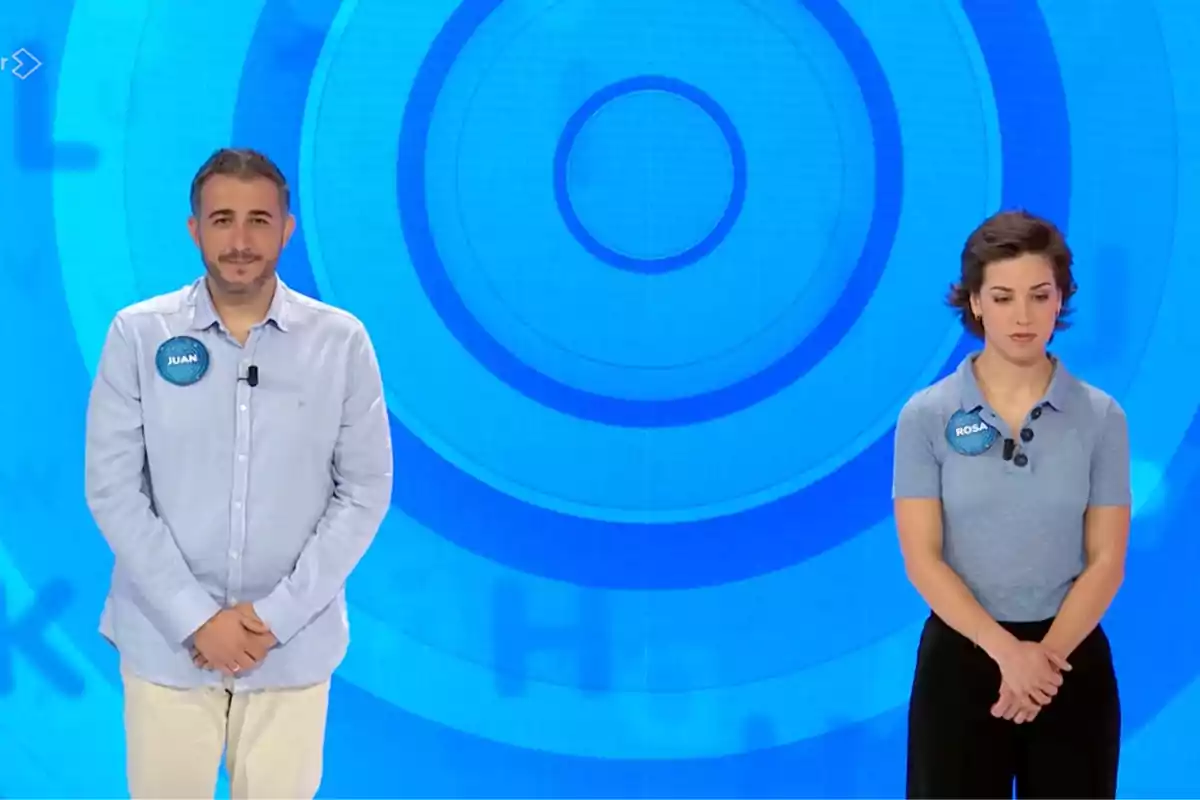 Joan i Rosa drets en un plató de televisió amb fons blau i cercles concèntrics, tots dos porten xapes amb els seus noms i miren al davant. Captura de 'Pasapalabra' Joan i Rosa drets en un plató de televisió amb fons blau i cercles concèntrics, tots dos porten xapes amb els seus noms i miren al davant. Captura de 'Pasapalabra'