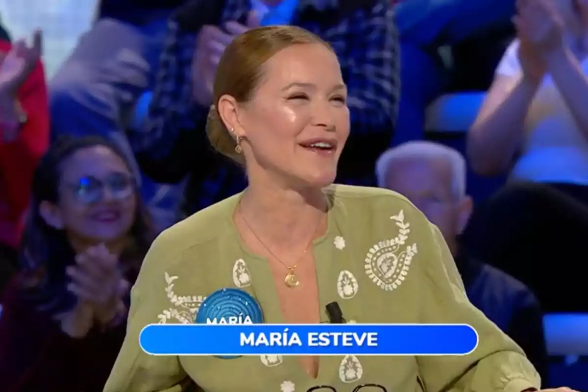 María Esteve somrient amb els cabells recollits i una brusa verd clar participant en un programa de televisió amb públic aplaudint al fons i un rètol blau amb el nom María Esteve. Captura de 'Pasapalabra'