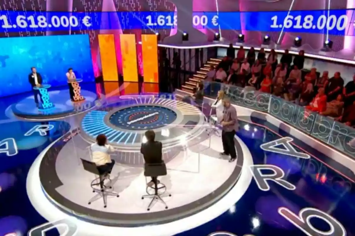 Un plató de concurs televisiu amb un disseny acolorit i modern, on s'observen participants en els seus respectius faristols, un presentador i una audiència asseguda al fons, amb una pantalla que mostra una quantitat de diners. Captura de 'Pasapalabra'. Un plató de concurs televisiu amb un disseny acolorit i modern, on s'observen participants en els seus respectius faristols, un presentador i una audiència asseguda al fons, amb una pantalla que mostra una quantitat de diners. Captura de 'Pasapalabra'.
