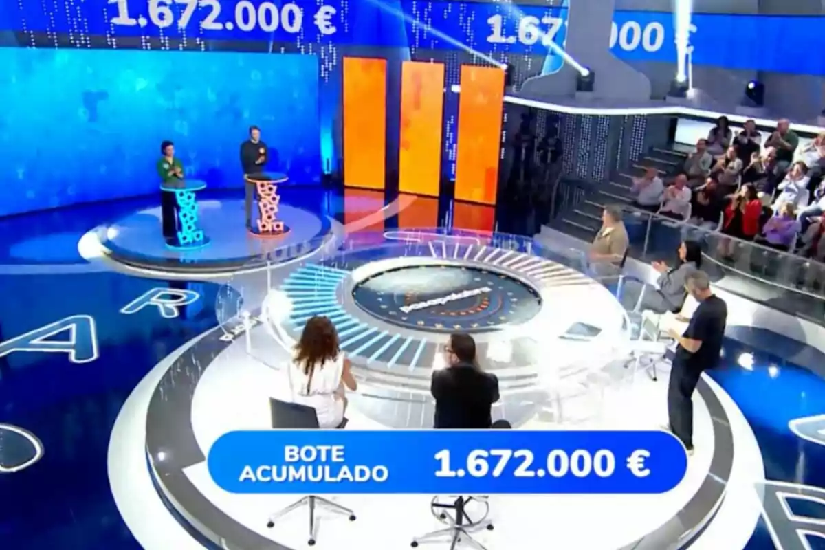 Escenari d’un concurs televisiu amb dos concursants drets a faristols il·luminats, un panell amb xifres grans en euros i públic observant des de les grades. Captura de ‘Pasapalabra’ Escenari d’un concurs televisiu amb dos concursants drets a faristols il·luminats, un panell amb xifres grans en euros i públic observant des de les grades. Captura de ‘Pasapalabra’