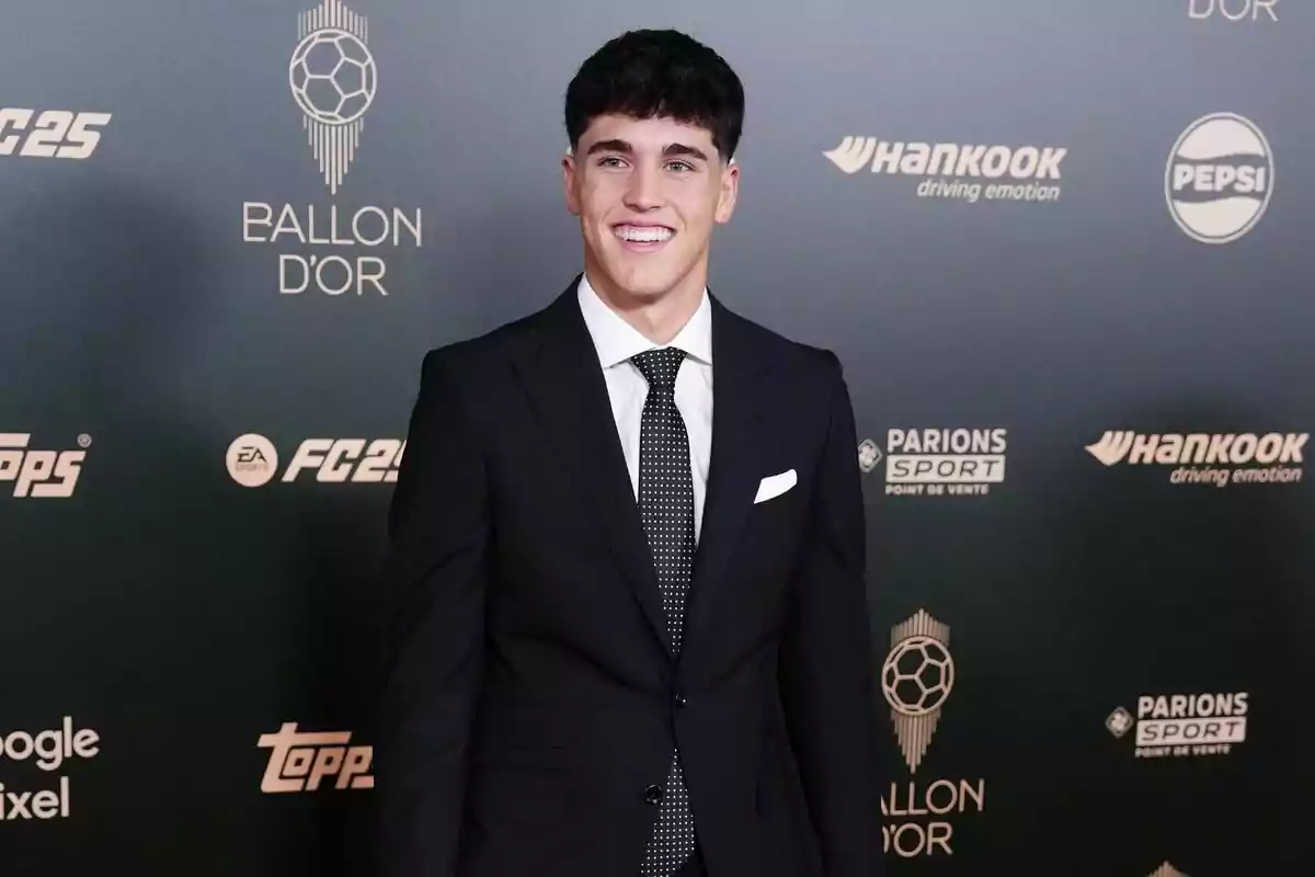 Un hombre joven con traje oscuro y corbata sonríe frente a un fondo con logotipos de marcas y el texto Un hombre joven con traje oscuro y corbata sonríe frente a un fondo con logotipos de marcas y el texto
