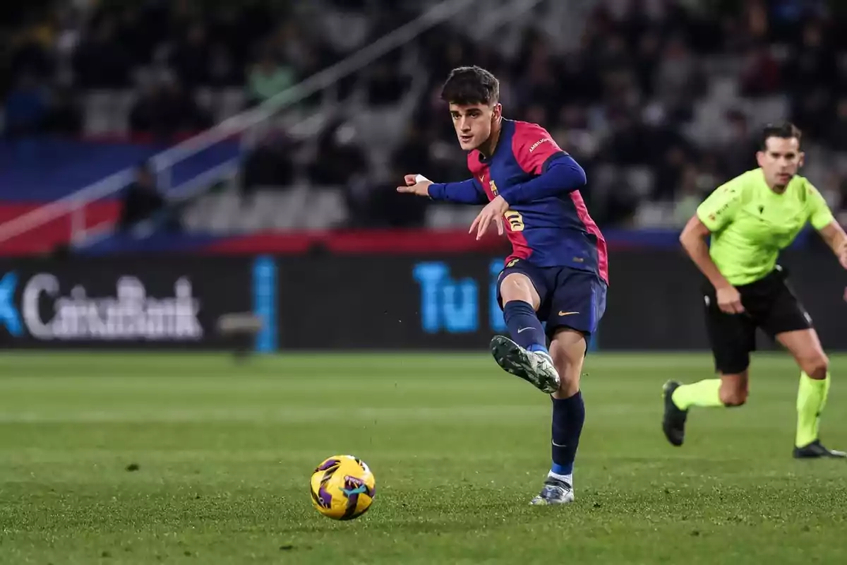 Un jugador de futbol amb uniforme blau i vermell pateja una pilota en un estadi mentre un àrbitre observa. Un jugador de futbol amb uniforme blau i vermell pateja una pilota en un estadi mentre un àrbitre observa.