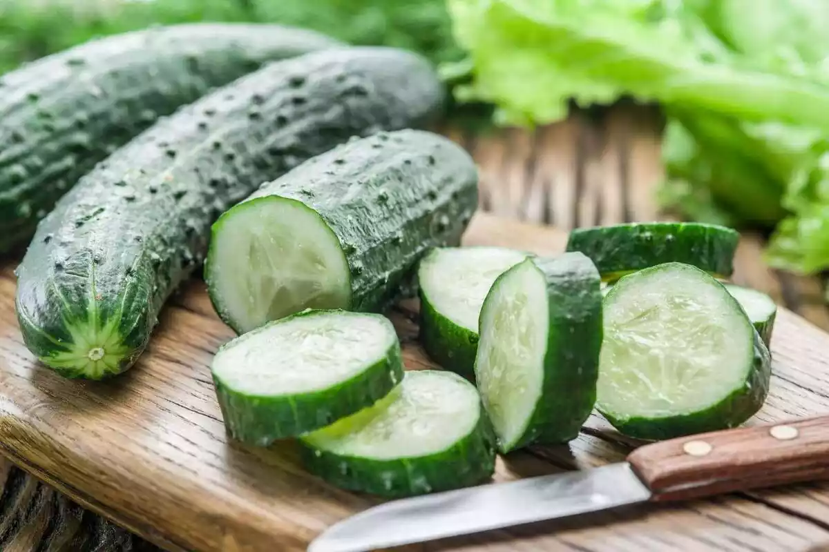 Dos cogombres sencers i un de tallat a rodanxes al costat d'un ganivet sobre un tauló de fusta Dos cogombres sencers i un de tallat a rodanxes al costat d'un ganivet sobre un tauló de fusta