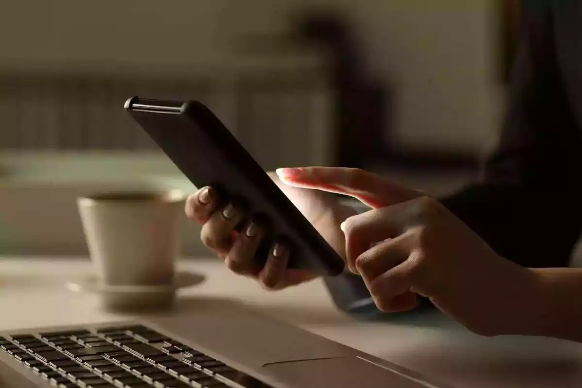 Una persona usant un mòbil amb un ordinador i una tassa de cafè al costat