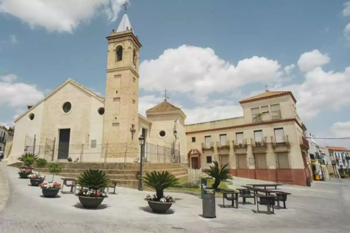 Una església de pedra amb una torre i un rellotge en una plaça amb bancs i testos sota un cel blau amb núvols. Una església de pedra amb una torre i un rellotge en una plaça amb bancs i testos sota un cel blau amb núvols.