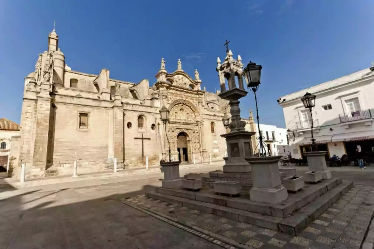 Església històrica amb façana detallada i plaça amb fanals i creu en primer pla sota un cel clar. Església històrica amb façana detallada i plaça amb fanals i creu en primer pla sota un cel clar.