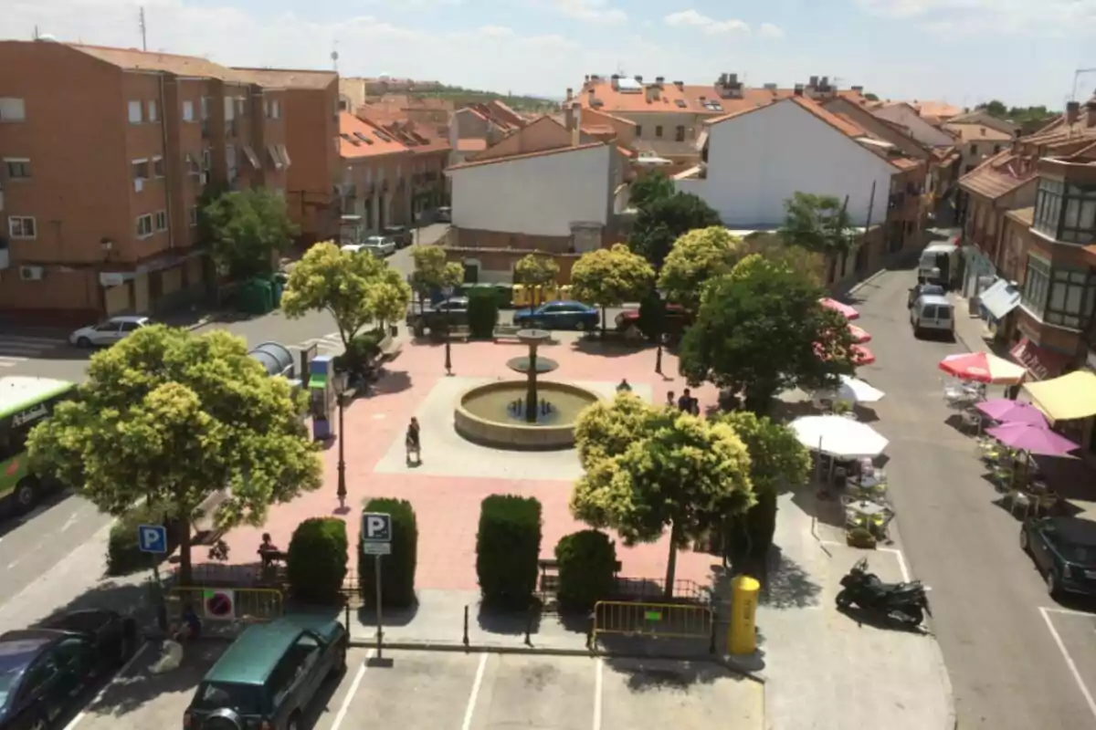 Una plaça amb una font central envoltada d'arbres i bancs, amb edificis de maó al voltant i diverses taules amb para-sols a la vorera. Una plaça amb una font central envoltada d'arbres i bancs, amb edificis de maó al voltant i diverses taules amb para-sols a la vorera.