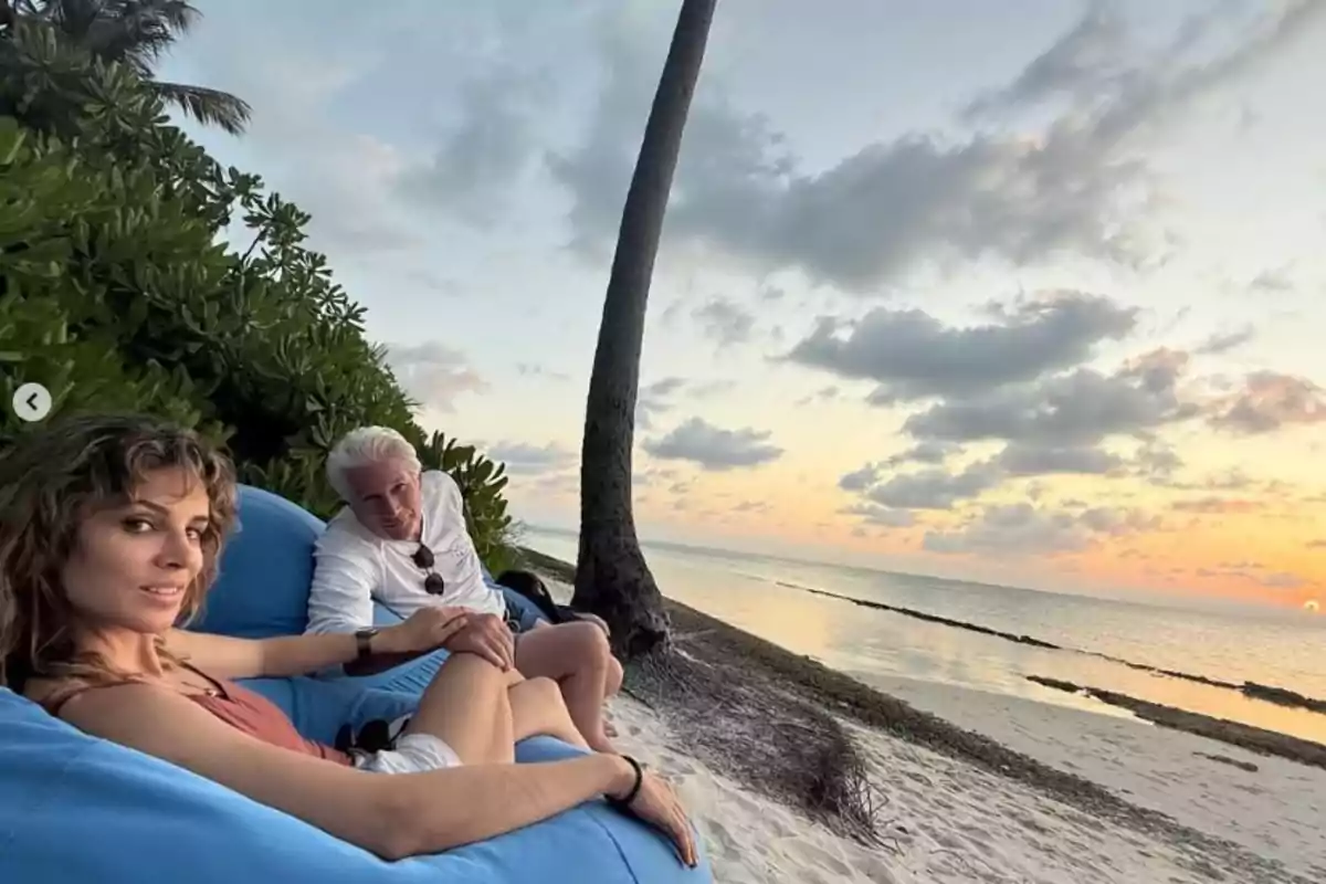 Alejandra Silva y Richard Gere relajándose en una playa al atardecer, sentados en sillones azules con el mar y el cielo nublado de fondo.