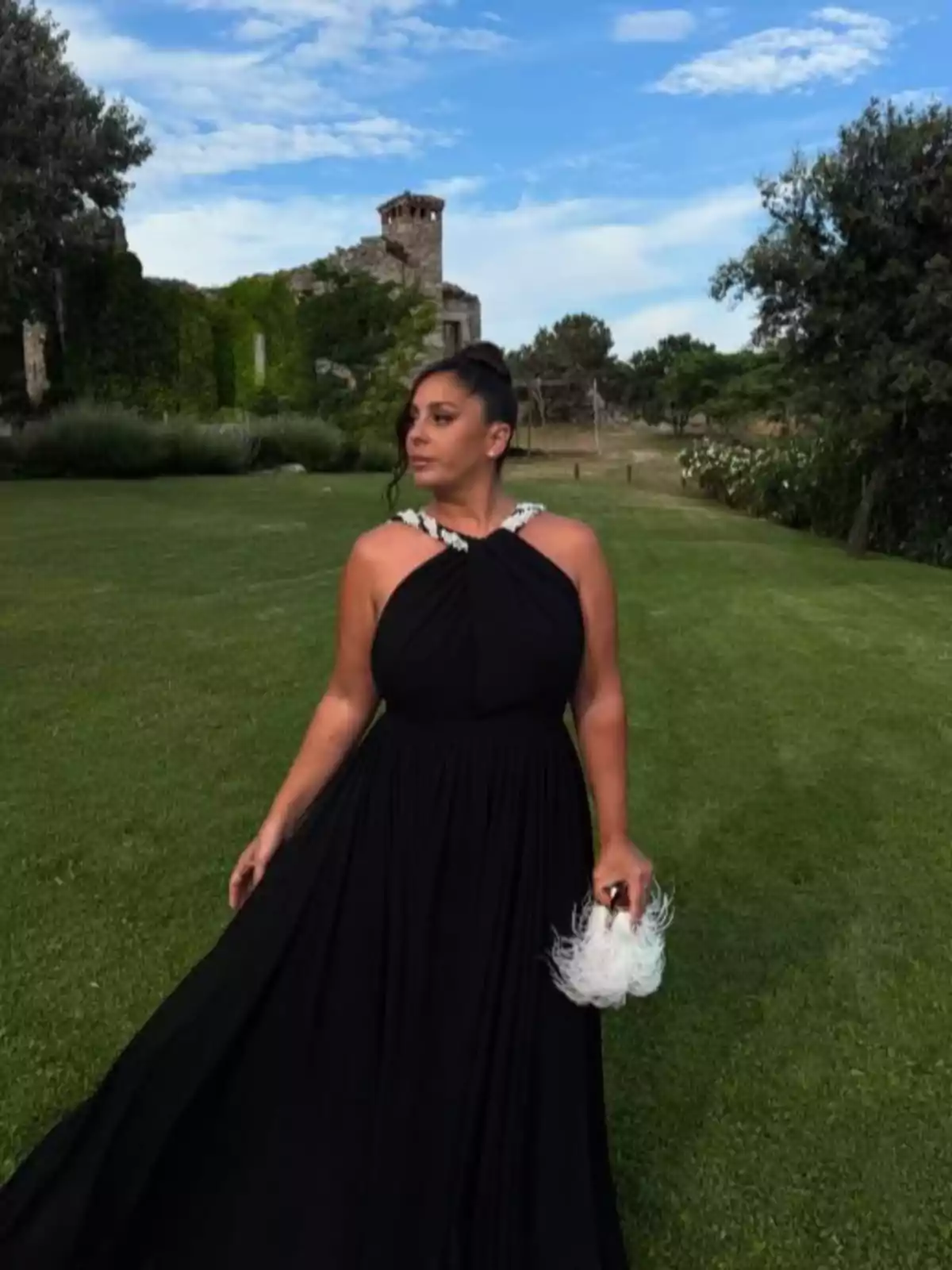 Anabel Pantoja con vestido largo negro y bolso blanco de plumas posando en un jardín con ruinas cubiertas de vegetación al fondo bajo un cielo azul con nubes Anabel Pantoja con vestido largo negro y bolso blanco de plumas posando en un jardín con ruinas cubiertas de vegetación al fondo bajo un cielo azul con nubes
