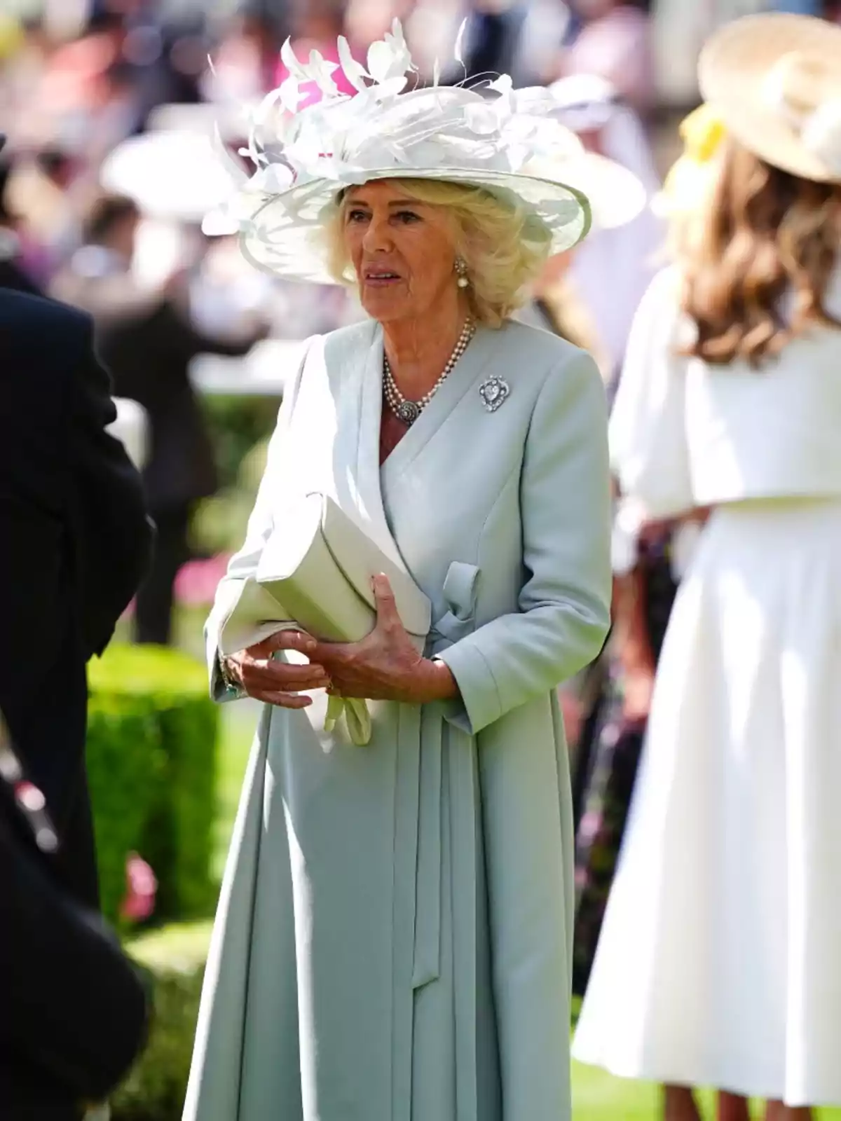 La reina Camila elegantment vestida amb un abric blau cel i un barret decorat amb flors blanques, sostenint una bossa en un esdeveniment a l’aire lliure. La reina Camila elegantment vestida amb un abric blau cel i un barret decorat amb flors blanques, sostenint una bossa en un esdeveniment a l’aire lliure.