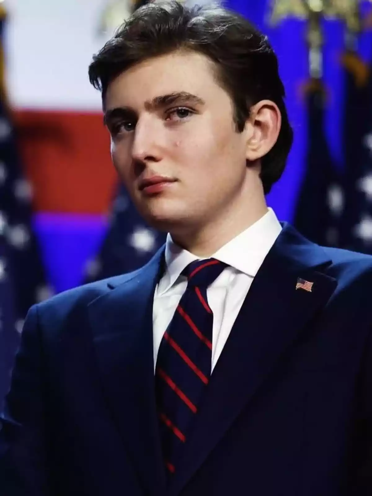 Barron Trump amb vestit blau i corbata de ratlles vermelles i blaves posant davant de banderes dels Estats Units Barron Trump amb vestit blau i corbata de ratlles vermelles i blaves posant davant de banderes dels Estats Units