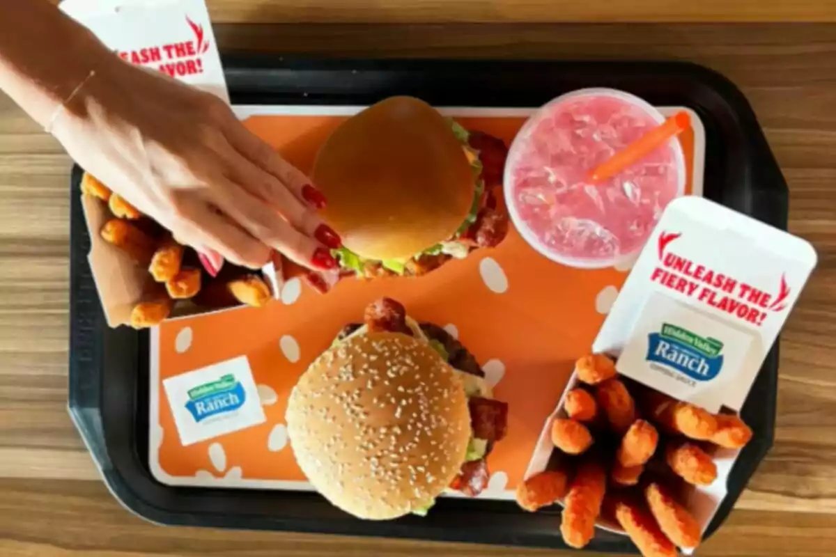 Una safata amb hamburgueses, patates fregides, una beguda rosada i un amaniment, amb una mà agafant una de les hamburgueses. Una safata amb hamburgueses, patates fregides, una beguda rosada i un amaniment, amb una mà agafant una de les hamburgueses.