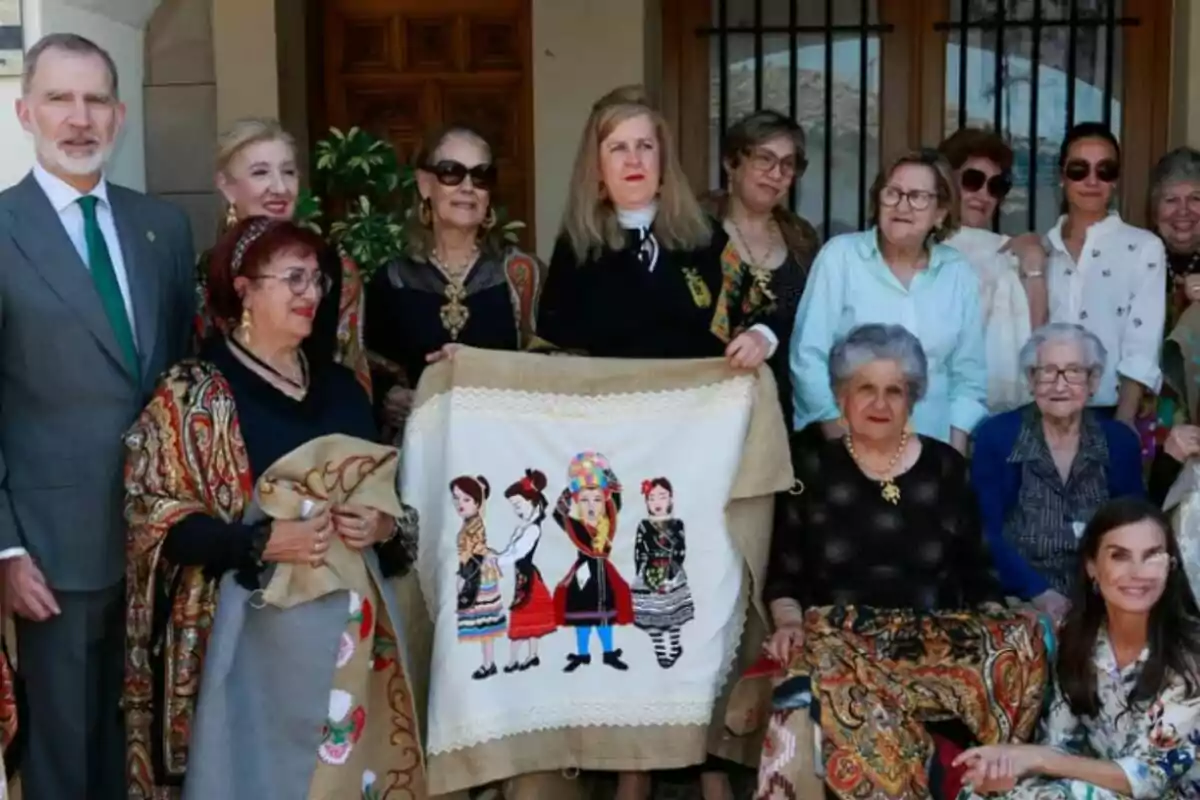 Group of people posing together, some wearing traditional clothing and a colorful embroidery with figures of women in the center Group of people posing together, some wearing traditional clothing and a colorful embroidery with figures of women in the center