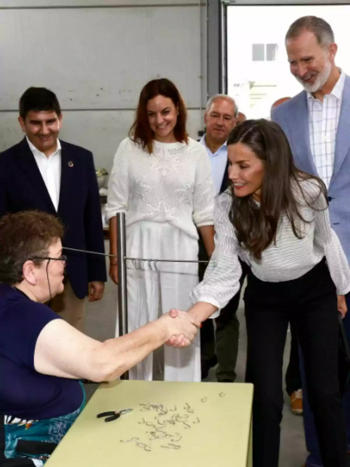 A woman sitting at a table shakes hands with Queen Letizia, who is standing, while several people watch and smile in an indoor setting. A woman sitting at a table shakes hands with Queen Letizia, who is standing, while several people watch and smile in an indoor setting.