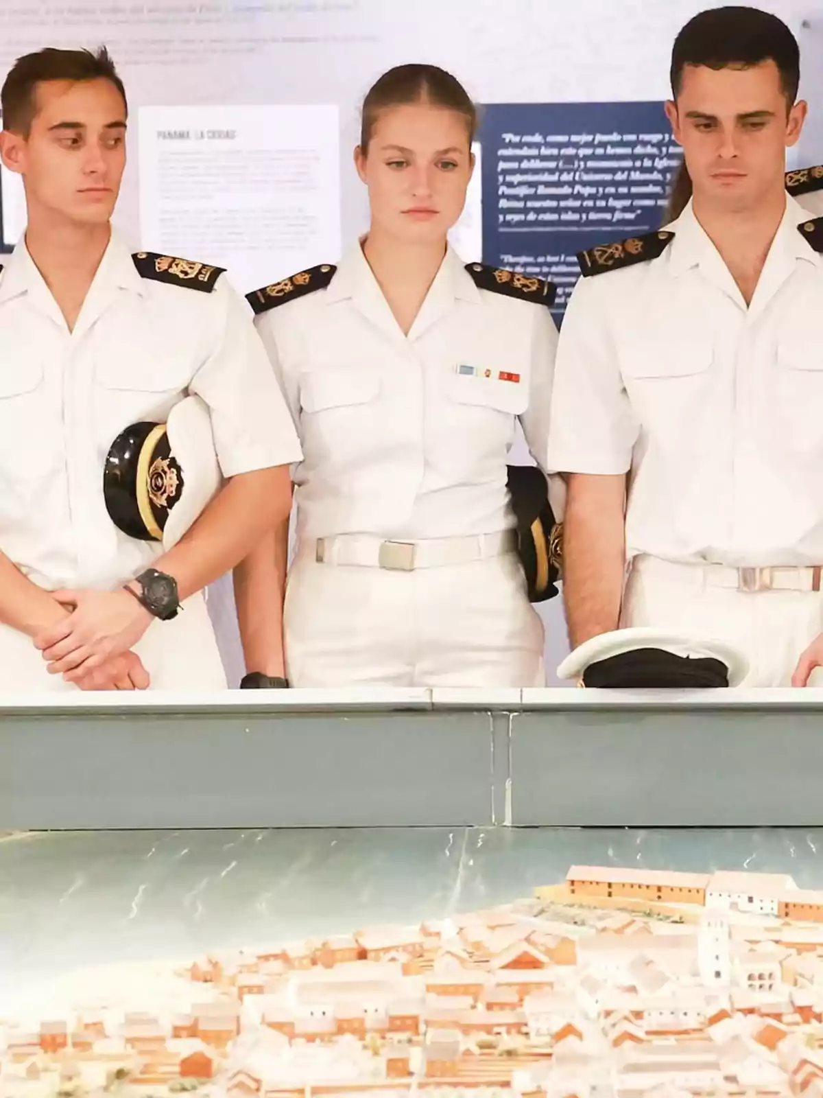 Tres persones vestides amb uniformes blancs d'estil naval observen un model a escala d'una ciutat. Tres persones vestides amb uniformes blancs d'estil naval observen un model a escala d'una ciutat.