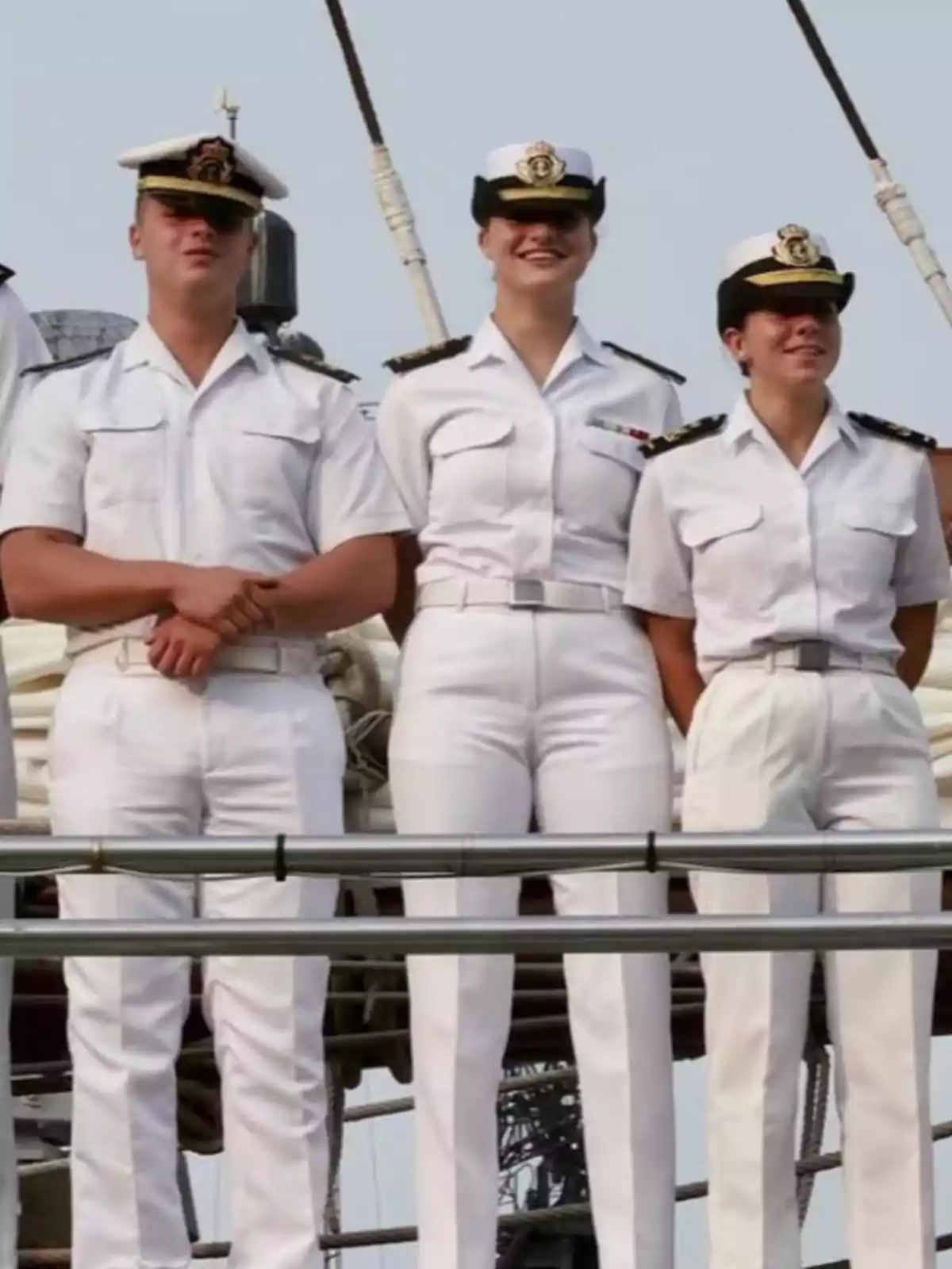 Princess Leonor and two companions in white navy uniforms stand on the deck of a ship. Princess Leonor and two companions in white navy uniforms stand on the deck of a ship.