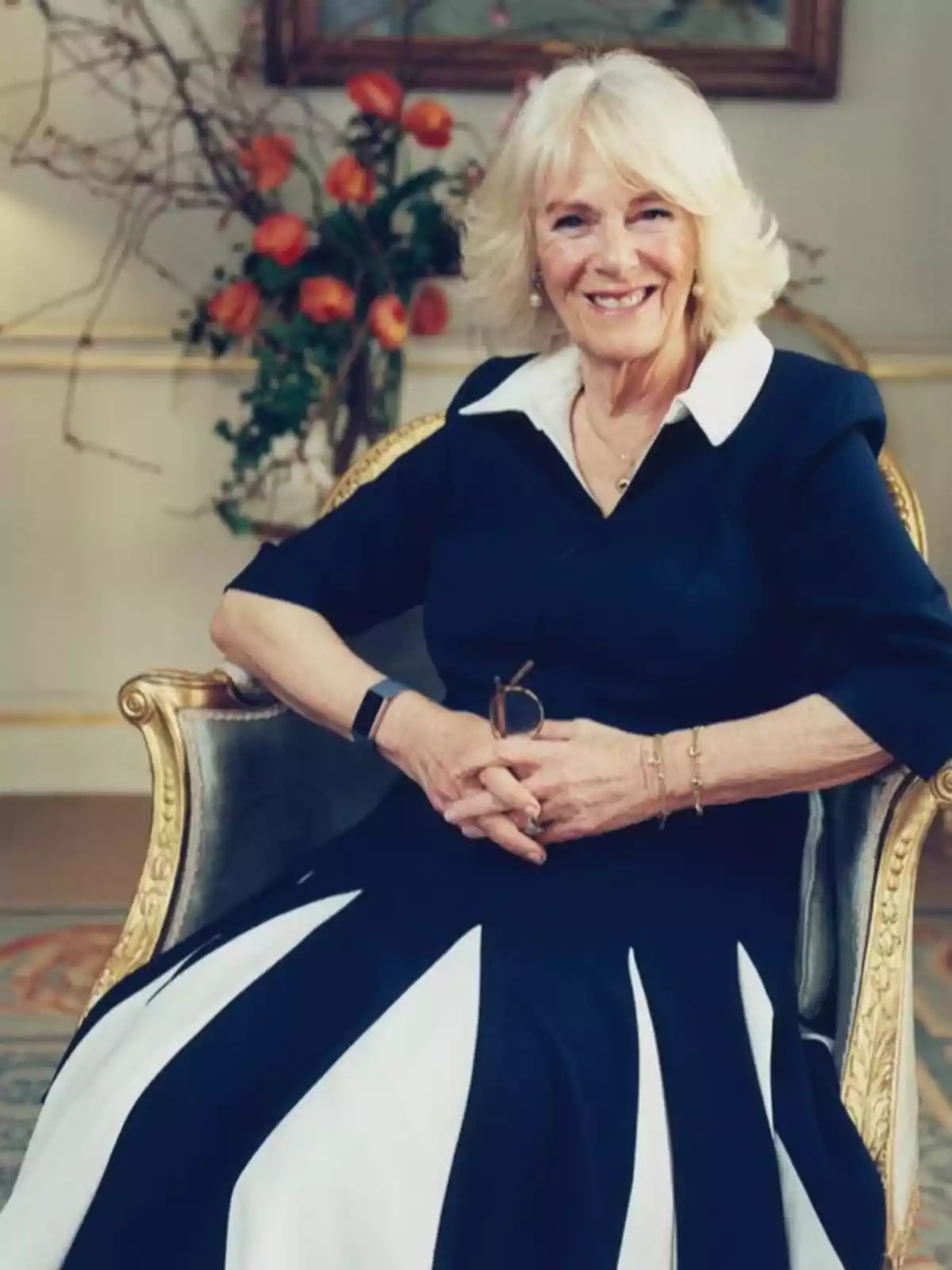 La reina Camila amb vestit blau fosc amb detalls blancs està asseguda en una cadira elegant, somrient davant d'un fons decorat amb flors taronges. La reina Camila amb vestit blau fosc amb detalls blancs està asseguda en una cadira elegant, somrient davant d'un fons decorat amb flors taronges.