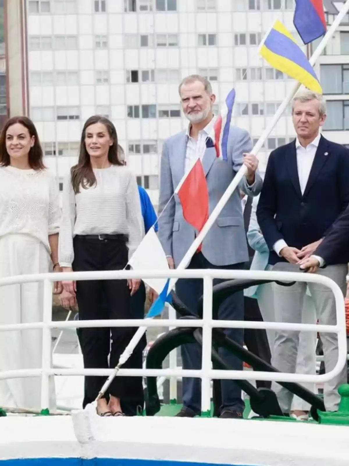 Queen Letizia, Felipe VI, and two other people pose standing on the deck of a boat with several colorful flags and a building in the background. Queen Letizia, Felipe VI, and two other people pose standing on the deck of a boat with several colorful flags and a building in the background.