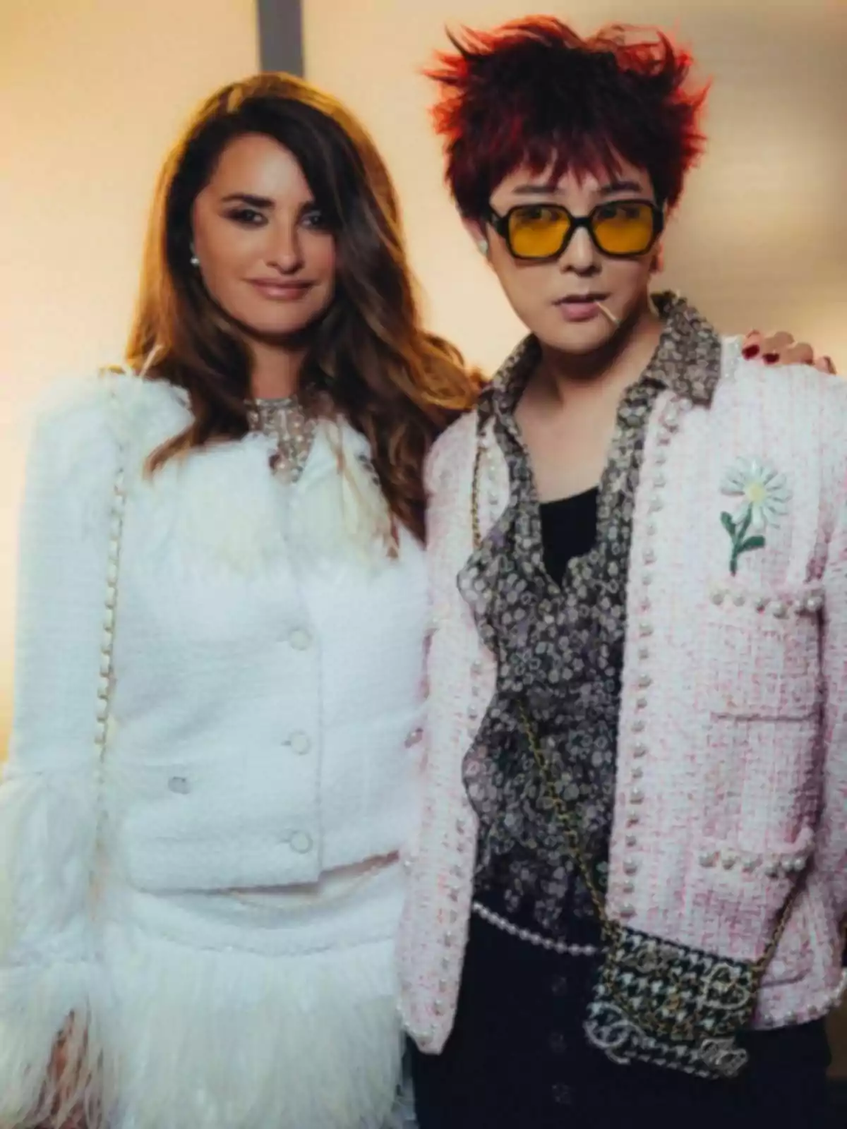 Two people pose together; one is wearing a white outfit with feather details and the other an outfit with a pink jacket, patterned jersey, and yellow sunglasses. Two people pose together; one is wearing a white outfit with feather details and the other an outfit with a pink jacket, patterned jersey, and yellow sunglasses.