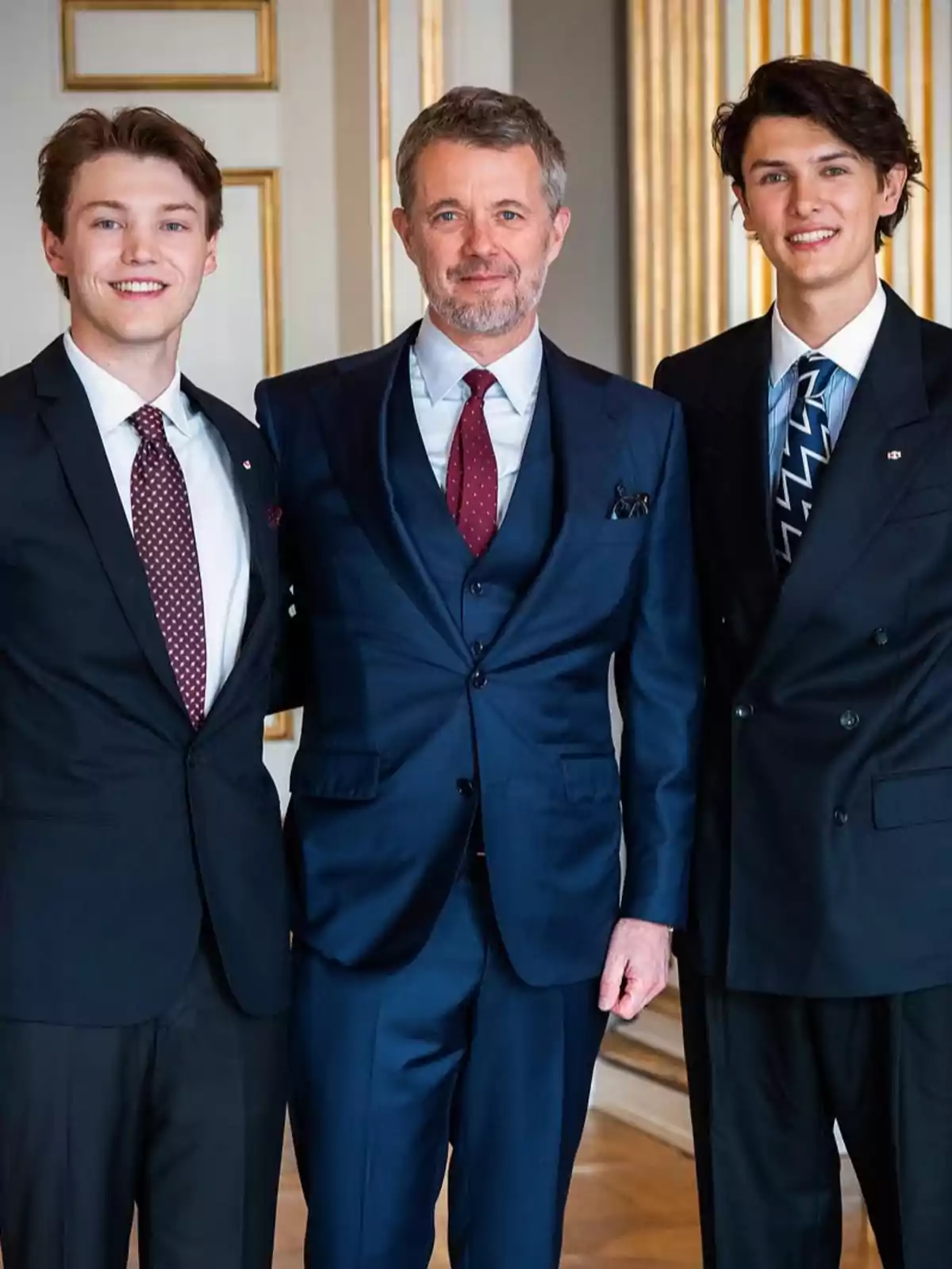Nikolai, Felix, and Frederik of Denmark, dressed in suits, pose together and smile in an elegant interior with gold details on the wall. Nikolai, Felix, and Frederik of Denmark, dressed in suits, pose together and smile in an elegant interior with gold details on the wall.