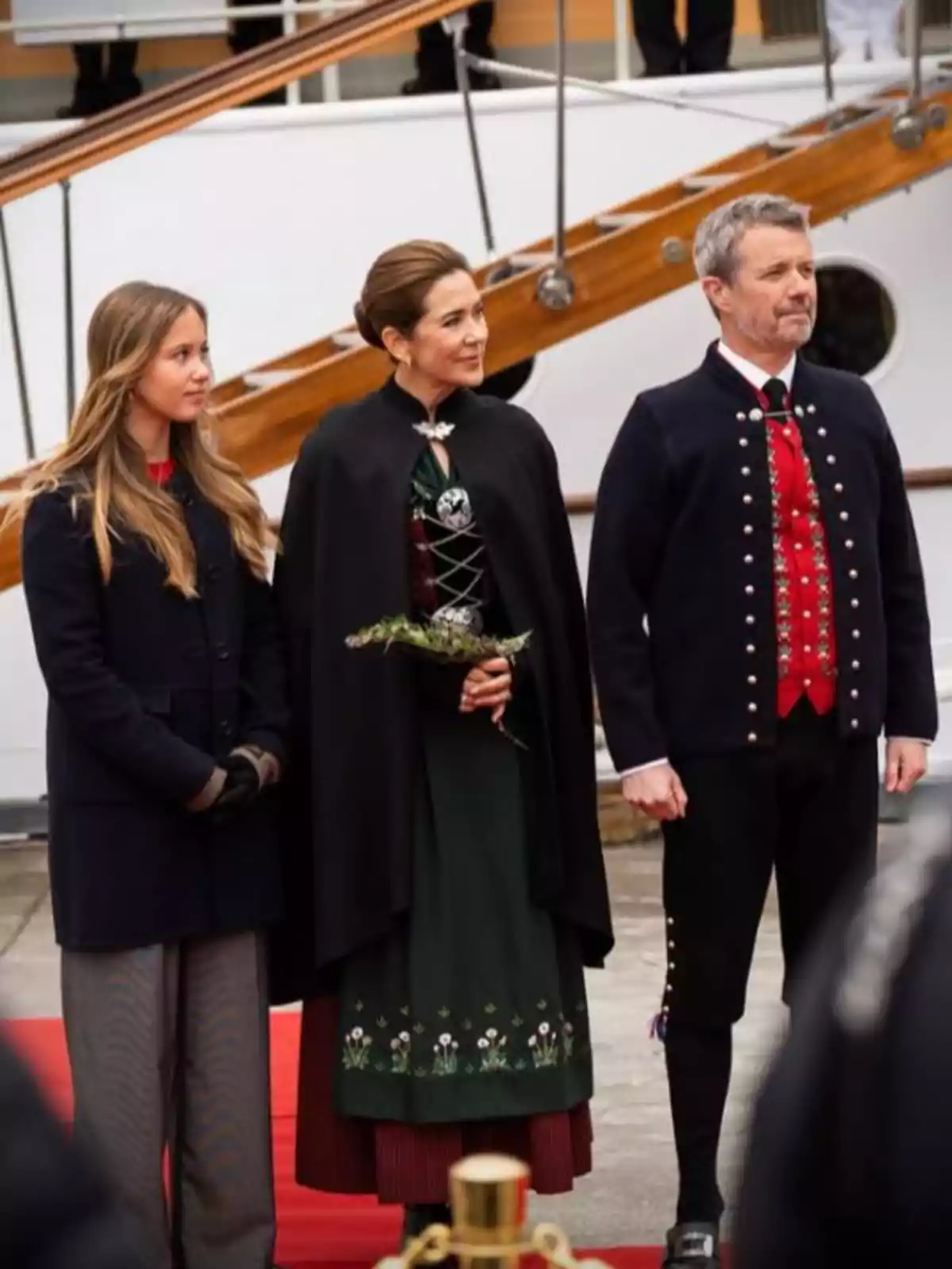 Josefina, Mary, and Frederik of Denmark standing on a red carpet, dressed in formal and traditional clothing, posing in front of a ship. Josefina, Mary, and Frederik of Denmark standing on a red carpet, dressed in formal and traditional clothing, posing in front of a ship.