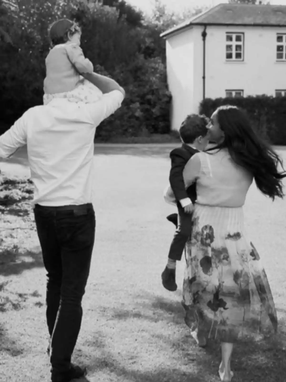 A family walking outdoors, with a man carrying a girl on his shoulders and a woman holding a boy in her arms, as they head toward a house. A family walking outdoors, with a man carrying a girl on his shoulders and a woman holding a boy in her arms, as they head toward a house.