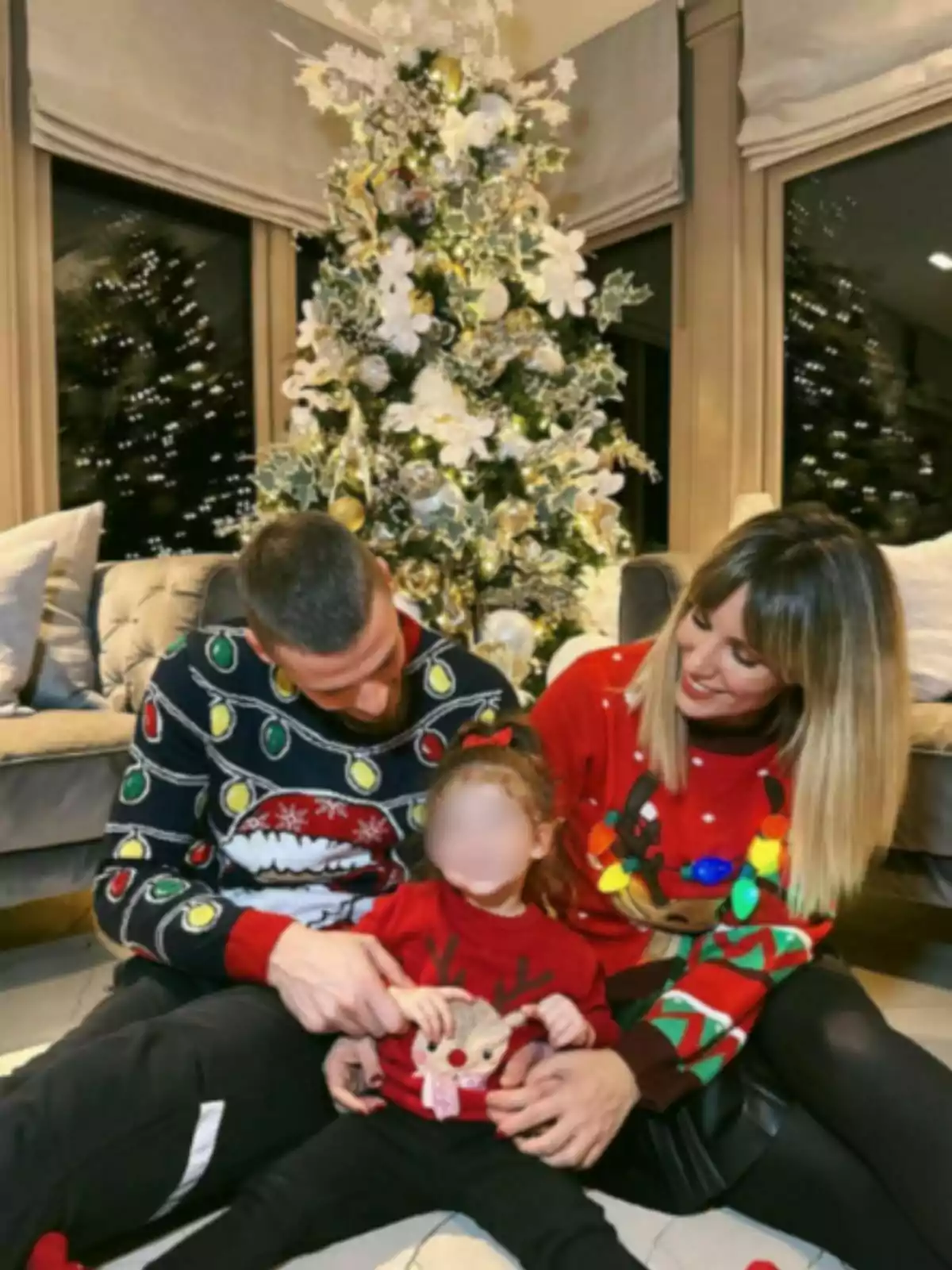 Família asseguda davant d’un arbre de Nadal decorat, portant jerseis nadalencs i somrient junts Família asseguda davant d’un arbre de Nadal decorat, portant jerseis nadalencs i somrient junts