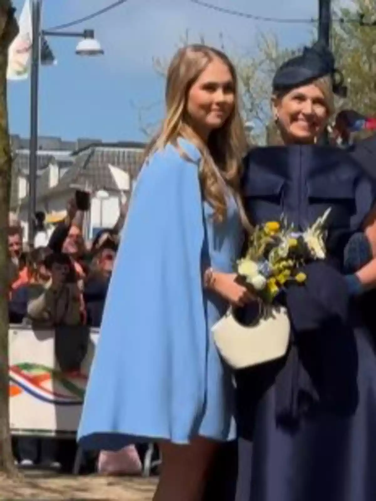 Dues dones posen juntes a l'aire lliure, una amb un vestit blau clar i l'altra amb una vestimenta blau fosc i un barret, mentre persones al fons fan fotos. Dues dones posen juntes a l'aire lliure, una amb un vestit blau clar i l'altra amb una vestimenta blau fosc i un barret, mentre persones al fons fan fotos.
