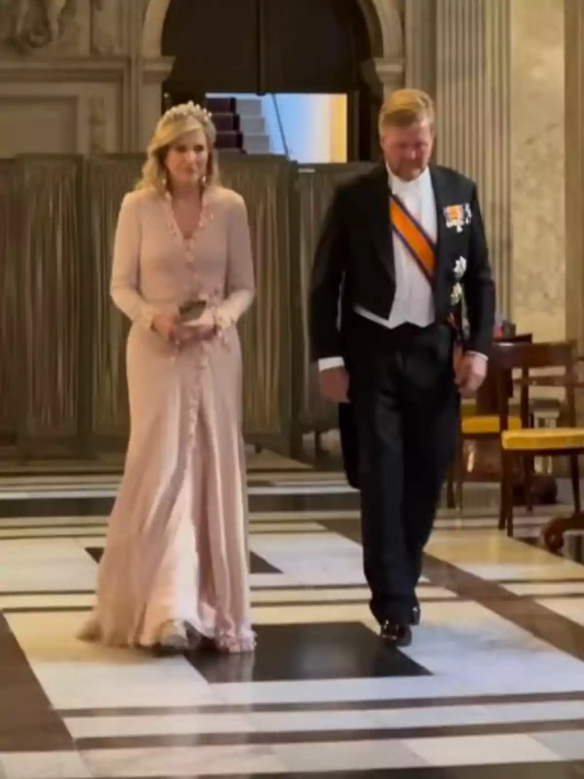 Máxima and Willem-Alexander of the Netherlands elegantly walk through a marble-floored hall; she wears a long dress and a tiara, while he is dressed in a formal suit with decorations. Máxima and Willem-Alexander of the Netherlands elegantly walk through a marble-floored hall; she wears a long dress and a tiara, while he is dressed in a formal suit with decorations.