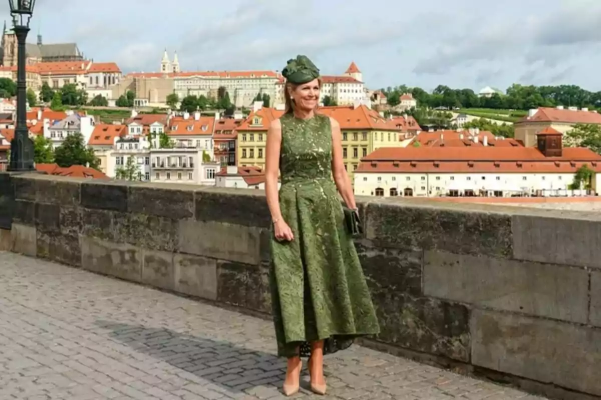 Màxima d’Holanda amb un vestit verd elegant i un barret a joc posant sobre un pont de pedra amb edificis de teulades vermelles i una ciutat històrica al fons Màxima d’Holanda amb un vestit verd elegant i un barret a joc posant sobre un pont de pedra amb edificis de teulades vermelles i una ciutat històrica al fons