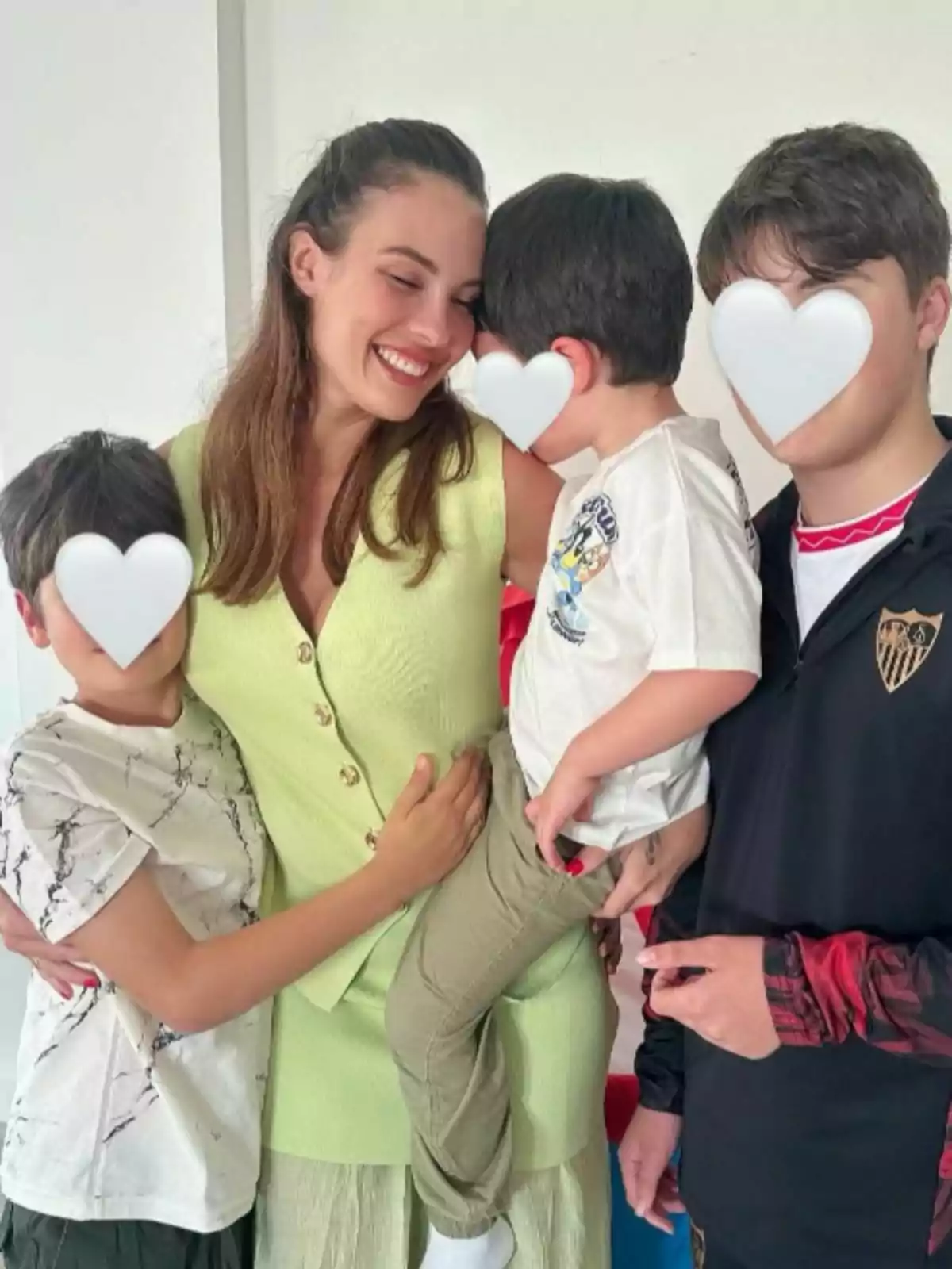 Jessica Bueno somrient sosté un nen petit mentre està envoltada d'altres dos nens, tots amb les cares cobertes per cors blancs. Jessica Bueno somrient sosté un nen petit mentre està envoltada d'altres dos nens, tots amb les cares cobertes per cors blancs.
