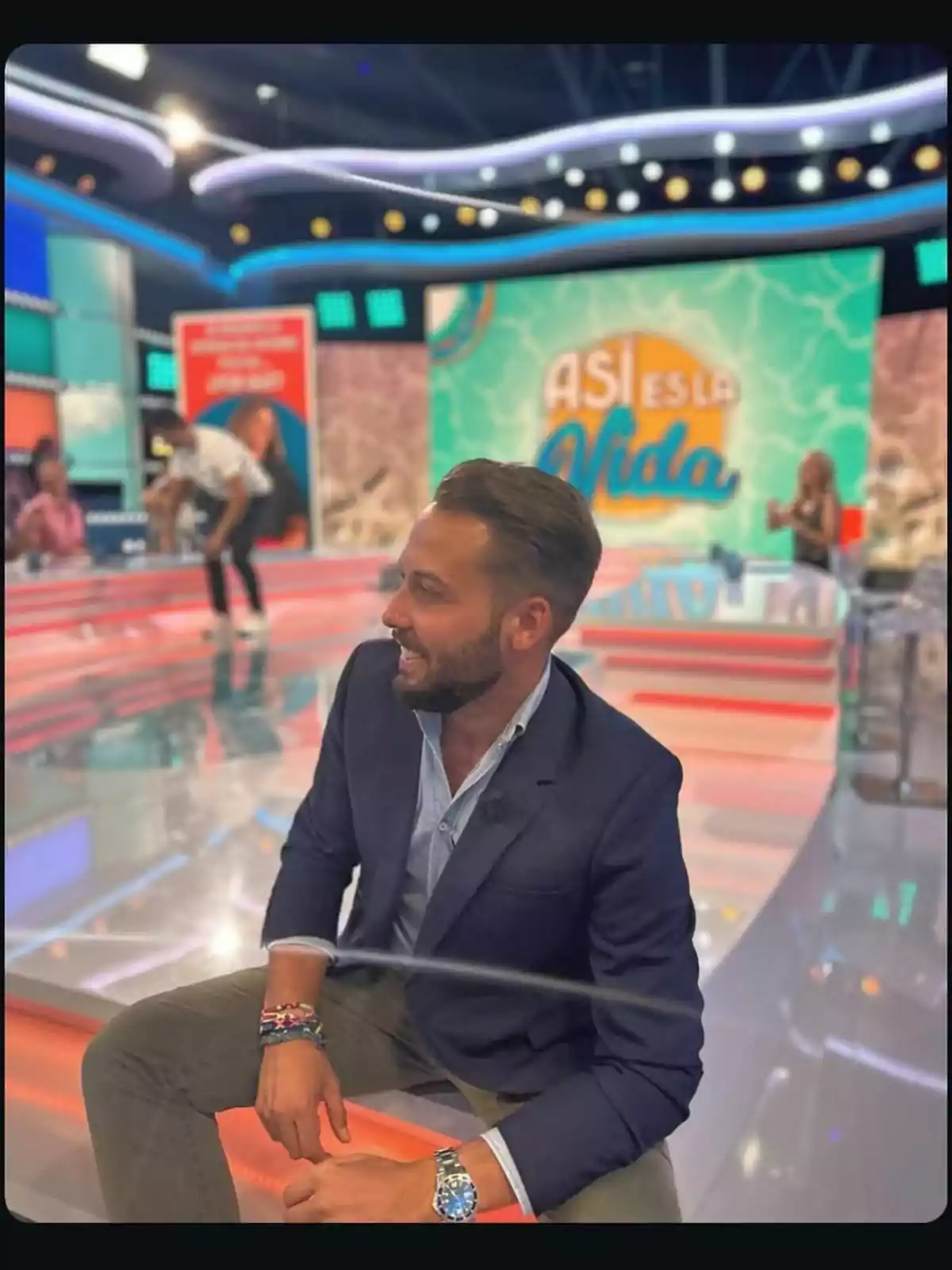 José Antonio Avilés sonriente está sentado en un set de televisión colorido con un letrero que dice José Antonio Avilés sonriente está sentado en un set de televisión colorido con un letrero que dice