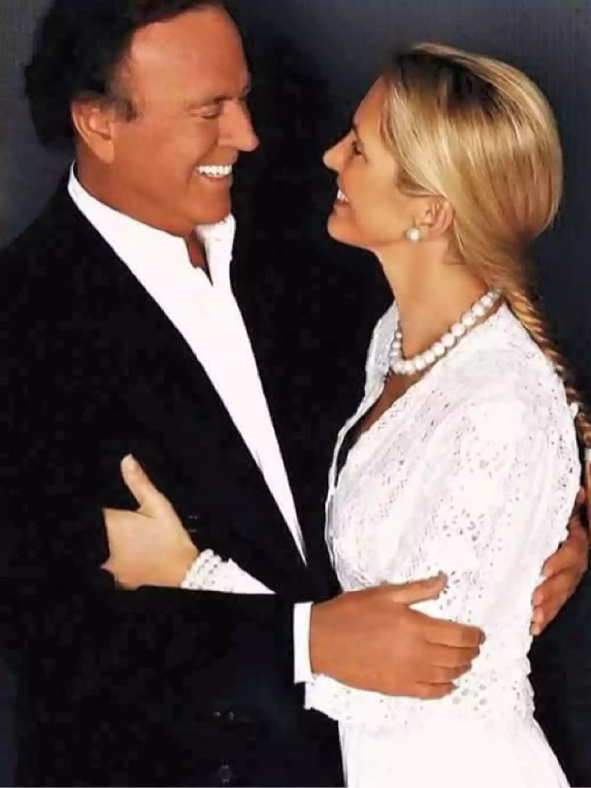 Julio Iglesias y Miranda Rijnsburger sonriente se mira a los ojos mientras se abraza, ambos visten de manera elegante con el hombre en traje oscuro y la mujer en un vestido blanco con perlas. Julio Iglesias y Miranda Rijnsburger sonriente se mira a los ojos mientras se abraza, ambos visten de manera elegante con el hombre en traje oscuro y la mujer en un vestido blanco con perlas.