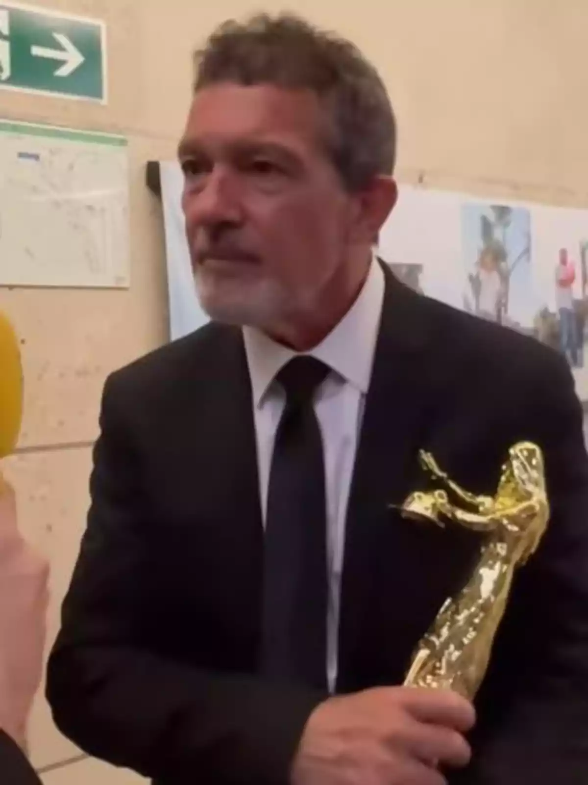 Antonio Banderas amb vestit fosc sosté un trofeu daurat mentre és entrevistat. Antonio Banderas amb vestit fosc sosté un trofeu daurat mentre és entrevistat.