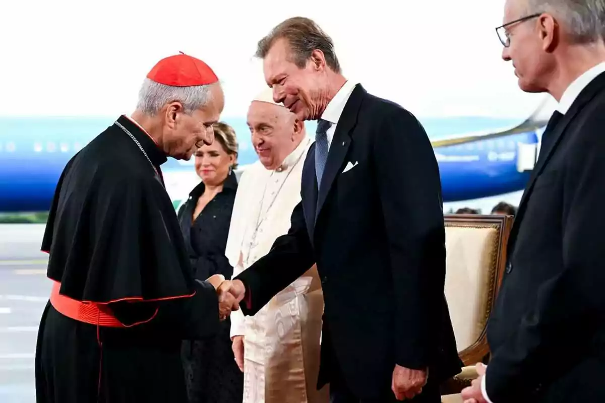A group of people, including a cardinal and a man in a suit, greet each other cordially in a formal setting with a plane in the background. A group of people, including a cardinal and a man in a suit, greet each other cordially in a formal setting with a plane in the background.