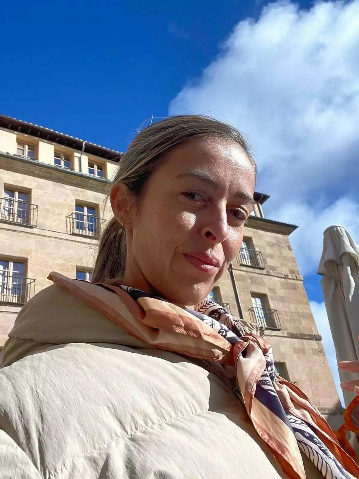 María Amores amb jaqueta clara i bufanda acolorida a l'aire lliure davant d'un edifici de pedra sota un cel blau amb núvols.