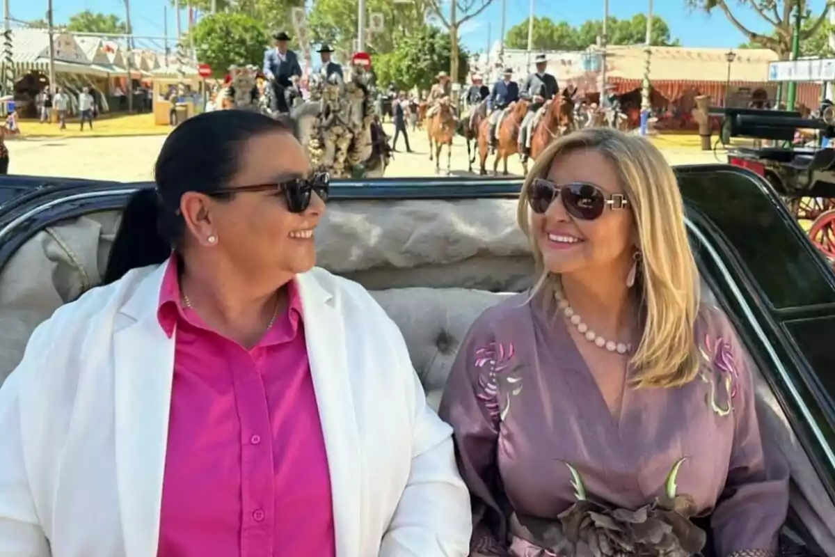 María del Monte i Inmaculada Casal somrients amb ulleres de sol estan assegudes en un carruatge durant un esdeveniment a l'aire lliure, amb persones a cavall i decoracions festives al fons. María del Monte i Inmaculada Casal somrients amb ulleres de sol estan assegudes en un carruatge durant un esdeveniment a l'aire lliure, amb persones a cavall i decoracions festives al fons.