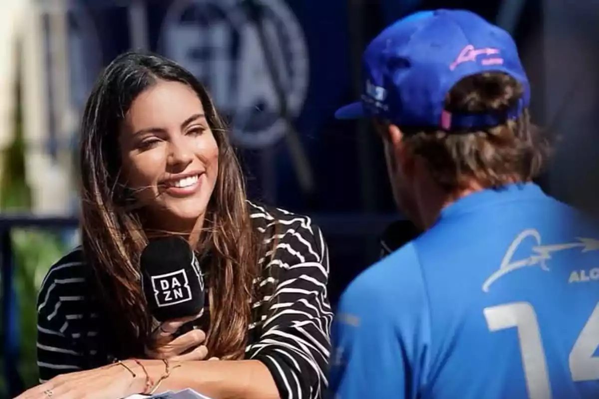 Malissa Jiménez sonriente sostiene un micrófono de DAZN mientras entrevista a Fernando Alonso con gorra y camiseta azul. Malissa Jiménez sonriente sostiene un micrófono de DAZN mientras entrevista a Fernando Alonso con gorra y camiseta azul.