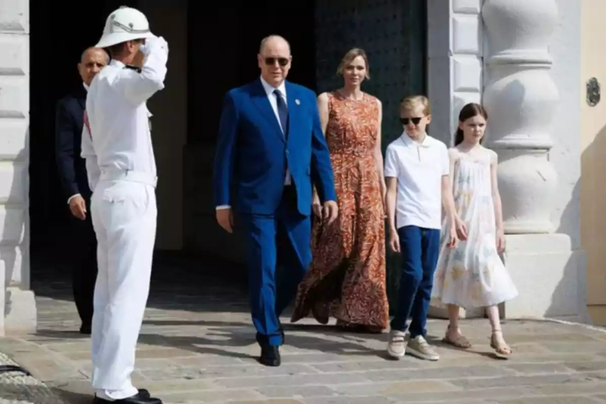 Una família elegantment vestida surt d’un edifici mentre un guàrdia amb uniforme blanc els saluda. Una família elegantment vestida surt d’un edifici mentre un guàrdia amb uniforme blanc els saluda.