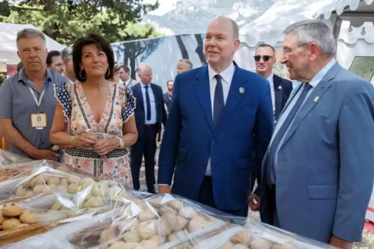 Un grupo de personas elegantemente vestidas observa una mesa llena de dulces y pasteles cubiertos con plástico en un evento al aire libre. Un grupo de personas elegantemente vestidas observa una mesa llena de dulces y pasteles cubiertos con plástico en un evento al aire libre.