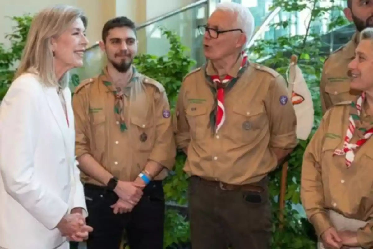 Un grup de persones vestides amb uniformes d'escoltes conversen amb una dona que porta un vestit blanc, en un entorn interior amb plantes. Un grup de persones vestides amb uniformes d'escoltes conversen amb una dona que porta un vestit blanc, en un entorn interior amb plantes.