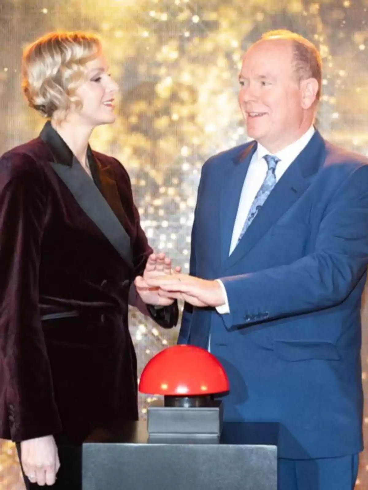 Charlene and Albert of Monaco, elegantly dressed, are together in front of a large red button at an event with a bright background. Charlene and Albert of Monaco, elegantly dressed, are together in front of a large red button at an event with a bright background.