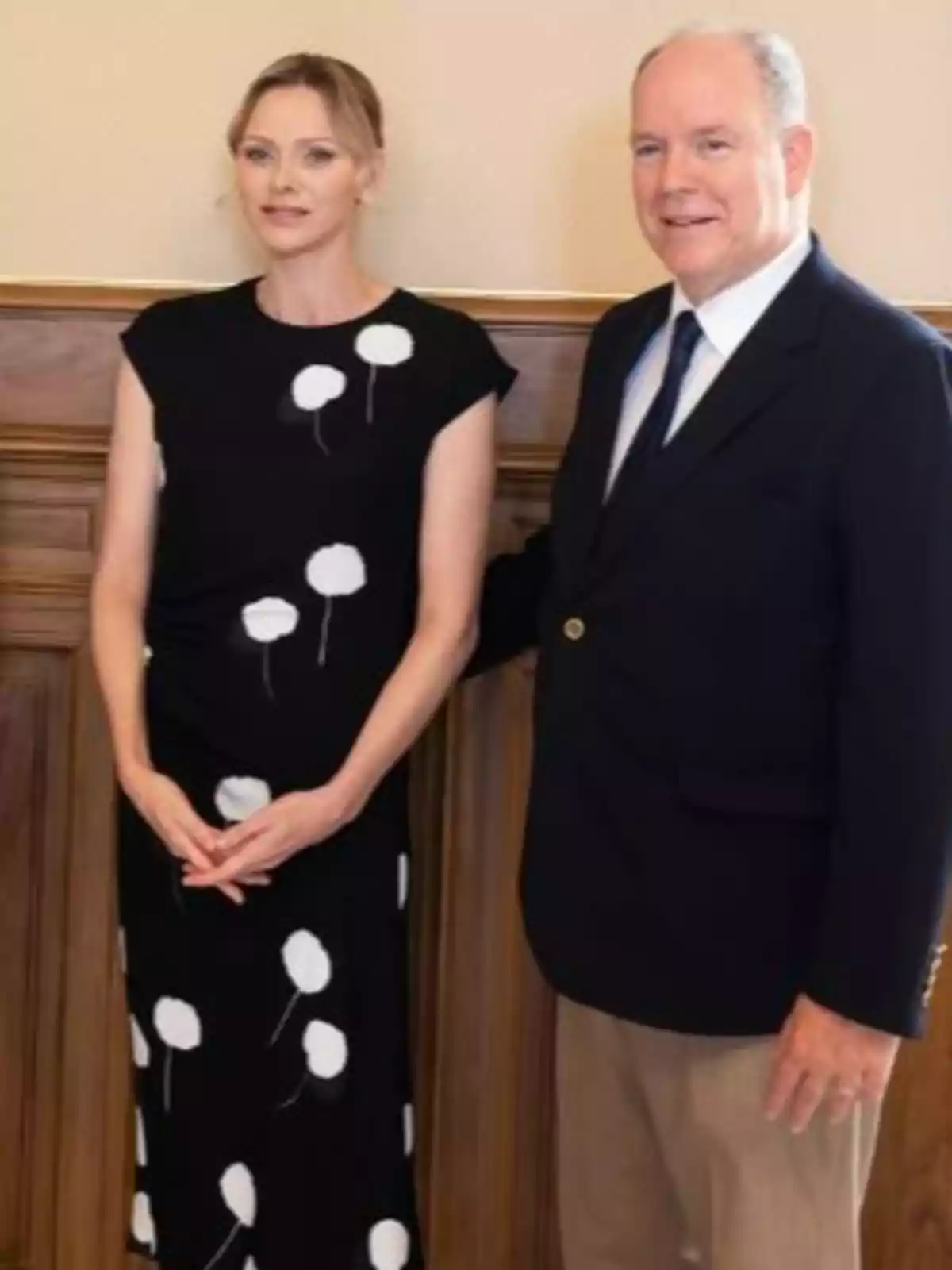 Charlene amb vestit negre de punts blancs i Albert de Mònaco amb americana fosca i pantalons clars posen junts en un interior elegant. Charlene amb vestit negre de punts blancs i Albert de Mònaco amb americana fosca i pantalons clars posen junts en un interior elegant.