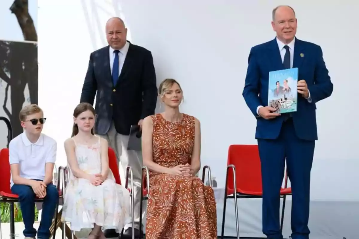 Cinco personas en un evento formal, dos niños sentados a la izquierda, una mujer con vestido largo sentada al centro, un hombre de traje azul de pie sosteniendo una revista y otro hombre de traje oscuro de pie al fondo Cinco personas en un evento formal, dos niños sentados a la izquierda, una mujer con vestido largo sentada al centro, un hombre de traje azul de pie sosteniendo una revista y otro hombre de traje oscuro de pie al fondo
