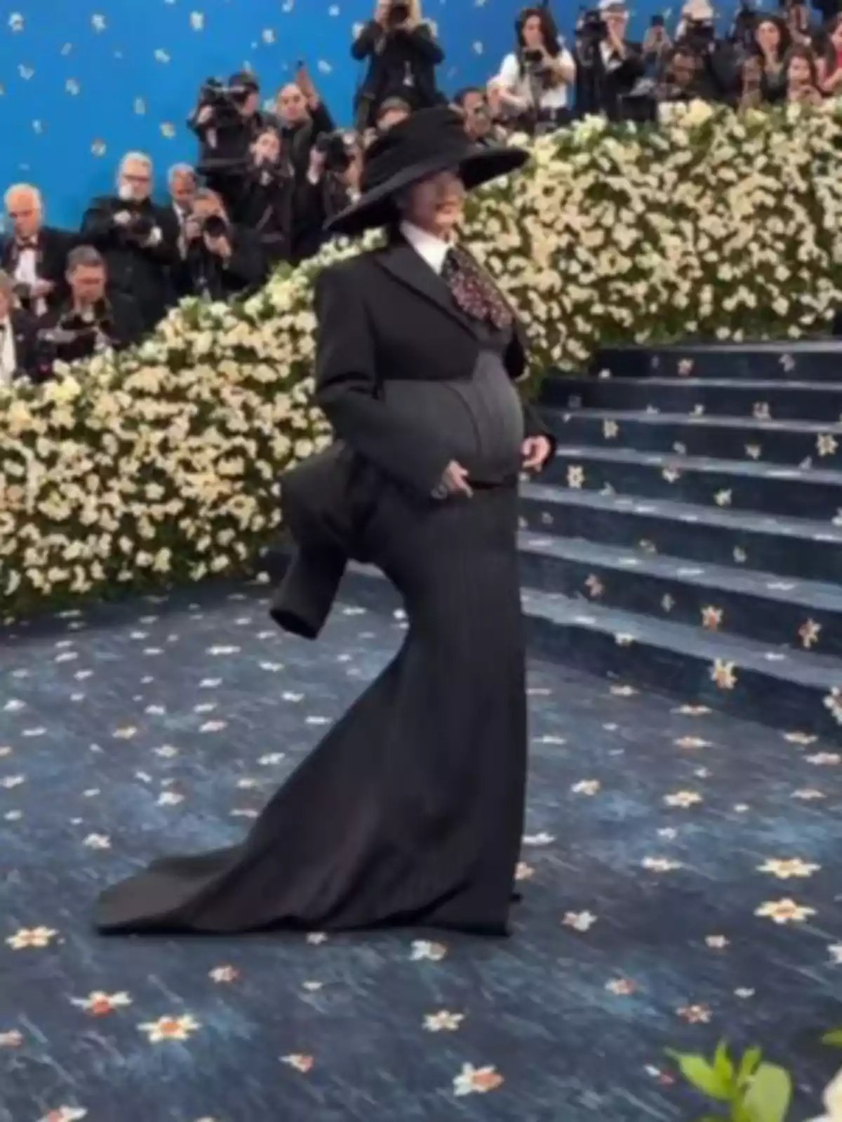 Rihanna in an extravagant black designer outfit walks on a blue carpet decorated with flowers, surrounded by photographers and a backdrop of white flowers. Rihanna in an extravagant black designer outfit walks on a blue carpet decorated with flowers, surrounded by photographers and a backdrop of white flowers.