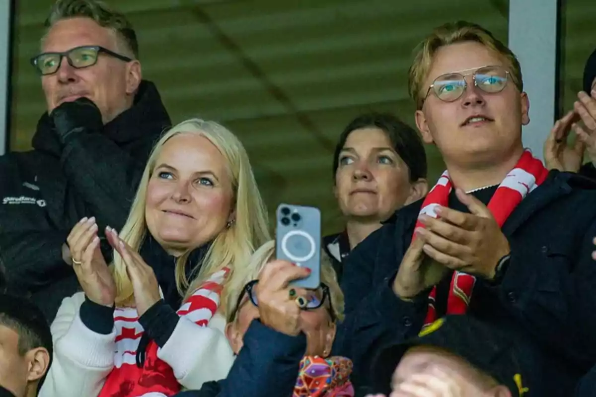 Un grupo de personas aplaude y observa algo fuera de la imagen, una mujer sostiene un teléfono móvil mientras otra lleva una bufanda roja y blanca. Un grupo de personas aplaude y observa algo fuera de la imagen, una mujer sostiene un teléfono móvil mientras otra lleva una bufanda roja y blanca.