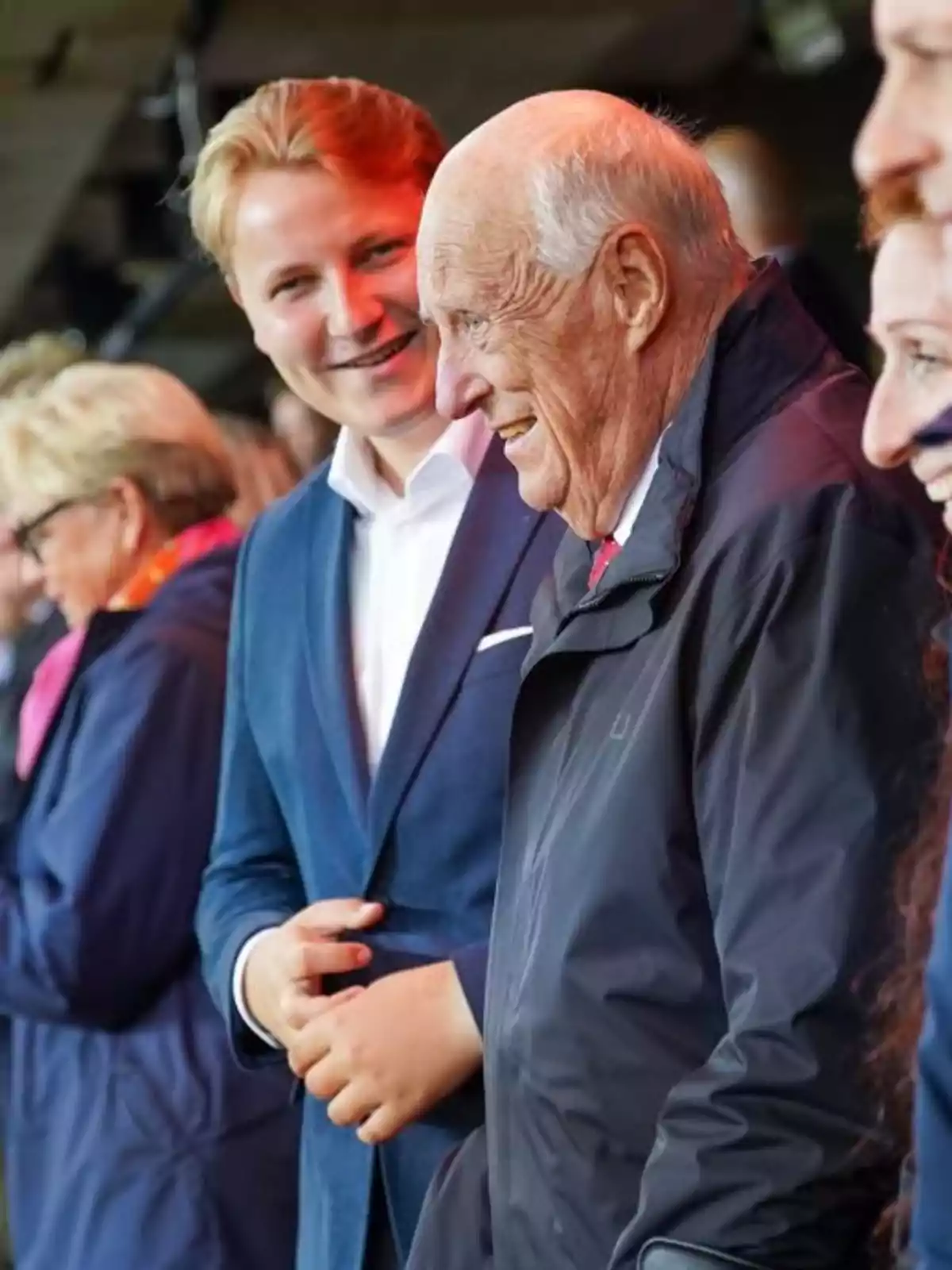 Smiling Harald of Norway in a dark jacket is accompanied by Sverre of Norway in a blue suit, both surrounded by other people in a social setting. Smiling Harald of Norway in a dark jacket is accompanied by Sverre of Norway in a blue suit, both surrounded by other people in a social setting.